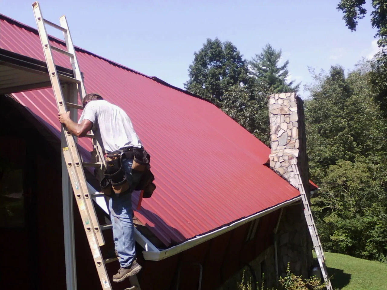 Roofer on a ladder beside a red metal roof and stone chimney — Soffit & Fascia Repair by Roofing Brothers in Troutville