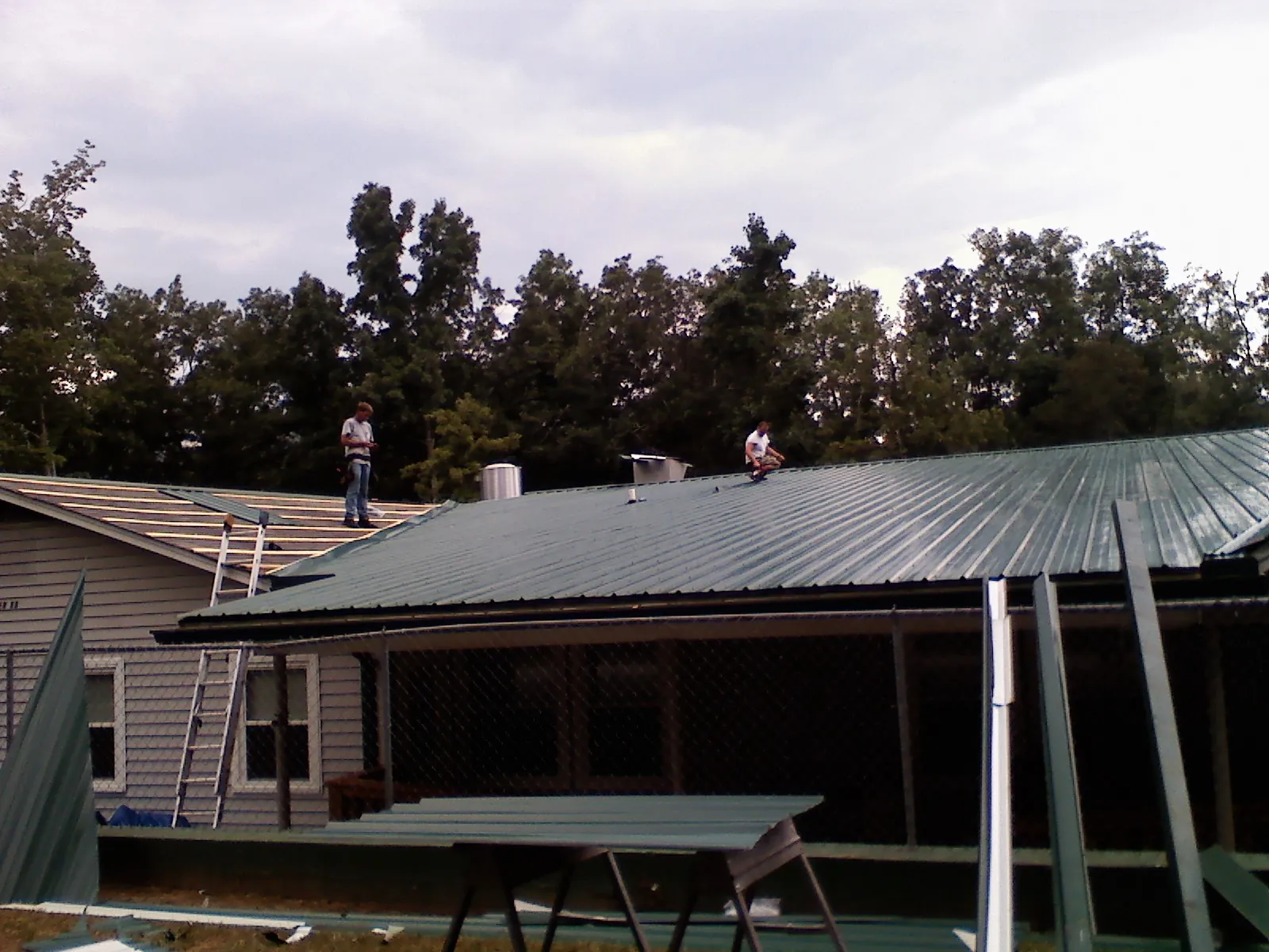 Two roofers installing green metal panels above a covered porch in Troutville